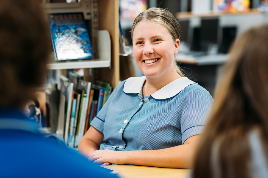 High school student sitting in library smiles to a friend.